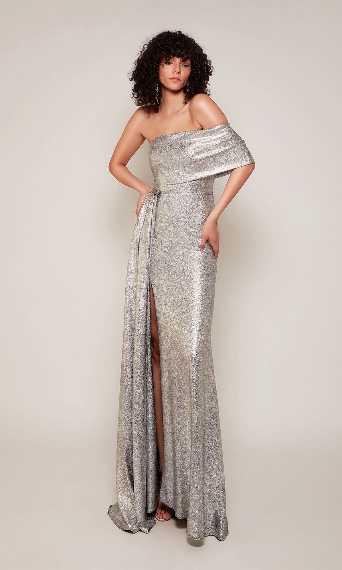 A person wears the silver Alyce 27663 long formal dress, featuring a metallic jacquard design, off-the-shoulder neckline, and thigh-high side slit, standing against a plain background.