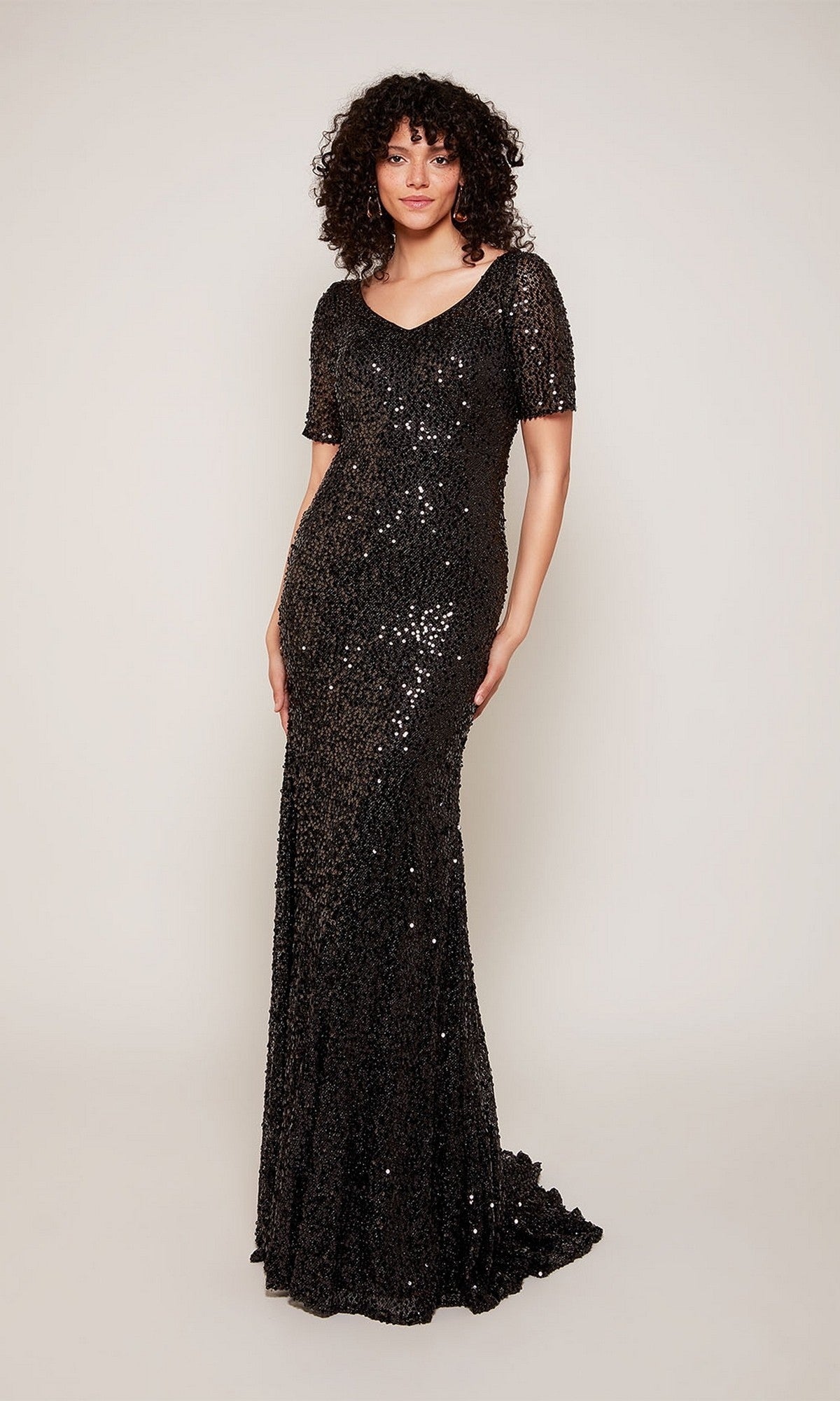 A woman with curly hair wears the Alyce 27668 long formal dress in black, featuring sequins, short sleeves, and a floor-length design—ideal for evening events. She stands against a plain light background.