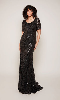 A woman with curly hair wears the Alyce 27668 long formal dress in black, featuring sequins, short sleeves, and a floor-length design—ideal for evening events. She stands against a plain light background.