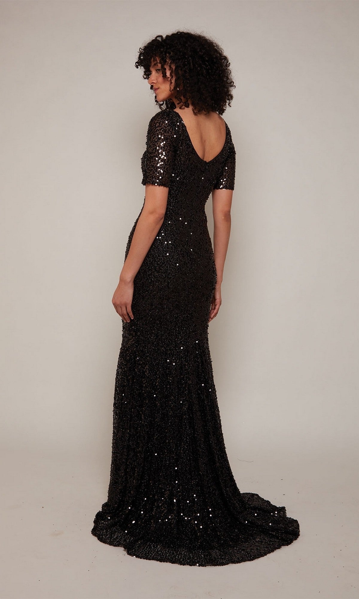 A woman with curly hair models the Alyce 27668 sequin long formal dress, showcasing short sleeves and a low back—an elegant choice for evening occasions against a plain light background.