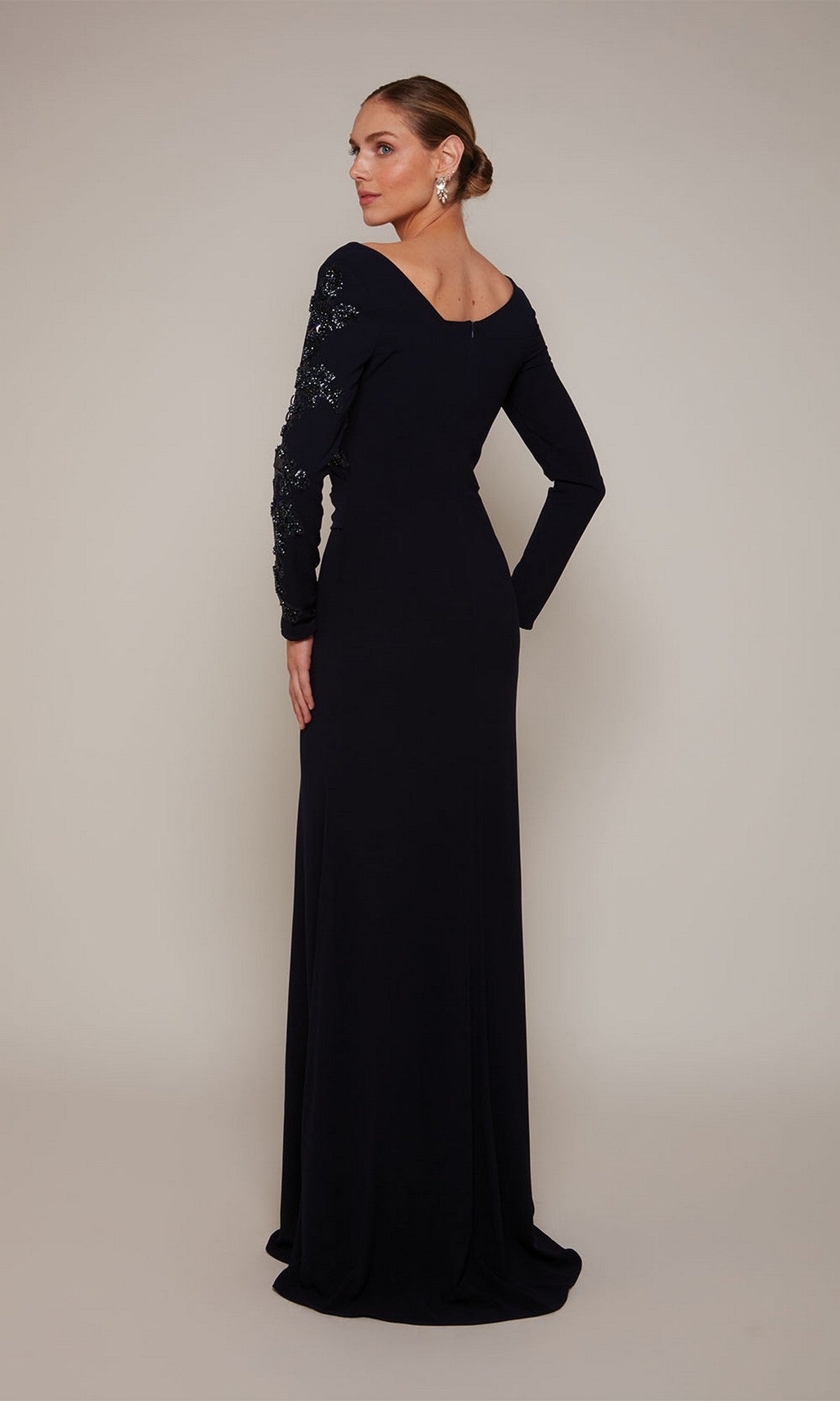 A woman, hair in a low bun, models the Alyce 27675 long formal dress with beaded detailing and one embellished sleeve, facing away against a plain background.