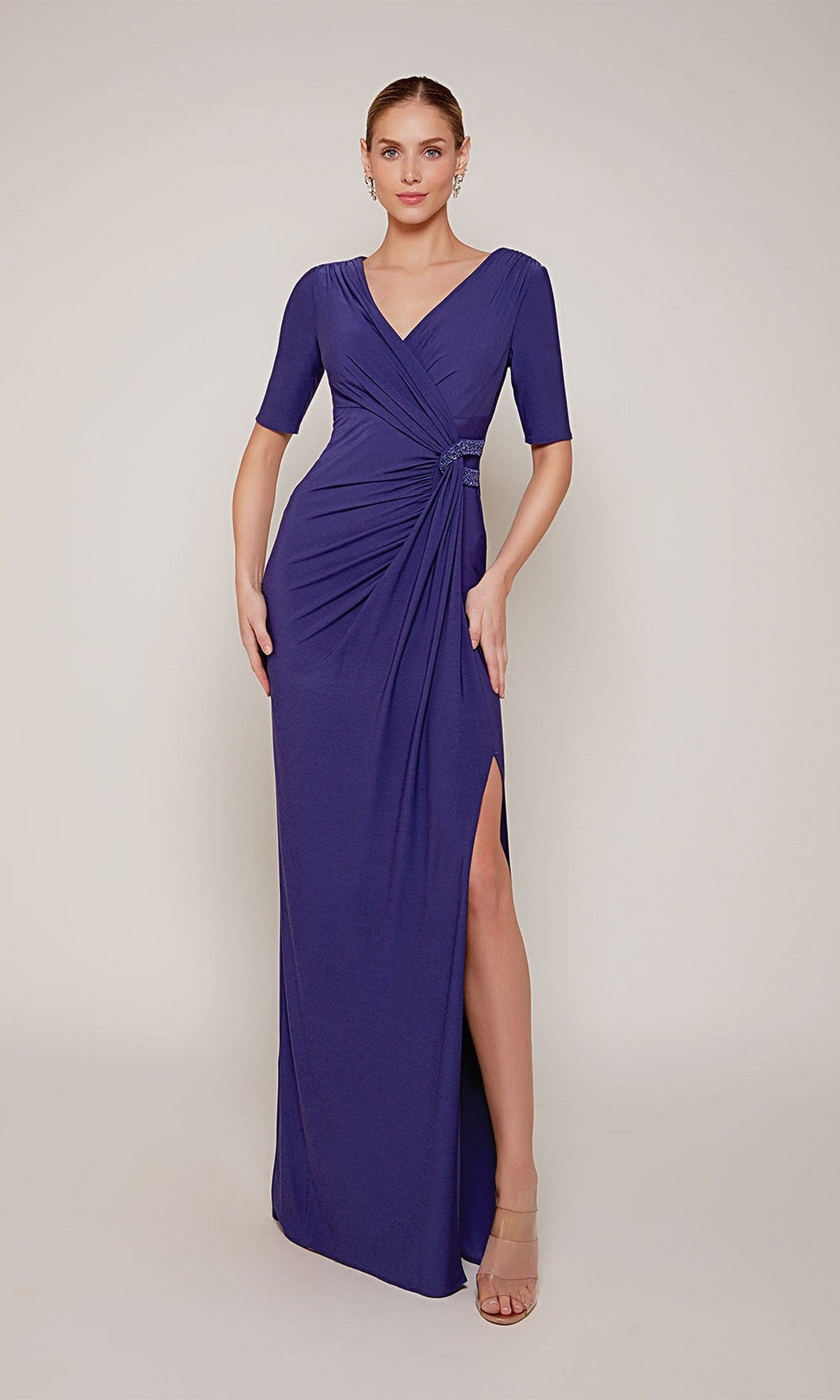 A woman stands in the Long Formal Dress: Alyce 27682 in royal, a floor-length gown with short sleeves, a v-neckline, side ruching, and a high slit. She pairs the formal jersey dress with nude heels against a plain background.