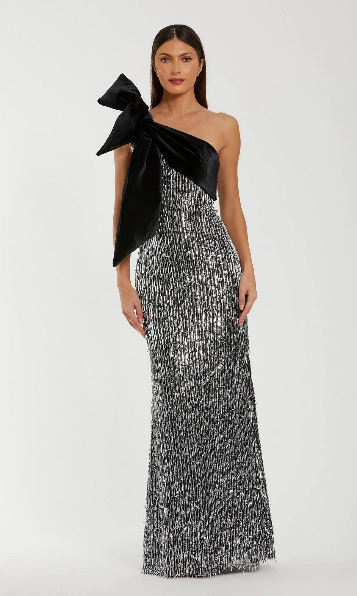 A woman wears the Mac Duggal 27690 Long Formal Dress, a one-shoulder sequin gown with a black velvet bow detail, standing against a plain light background.