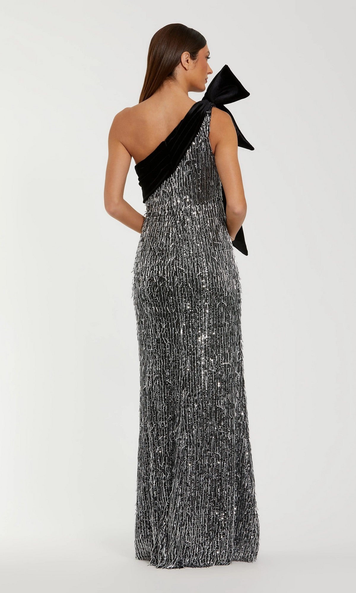 A woman faces away, wearing the Mac Duggal 27690 Long Formal Dress— a black and silver floor-length sequin gown with one shoulder and a dramatic bow detail.