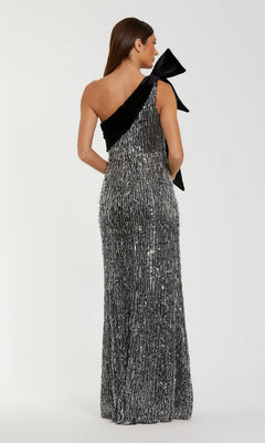 A woman faces away, wearing the Mac Duggal 27690 Long Formal Dress— a black and silver floor-length sequin gown with one shoulder and a dramatic bow detail.