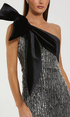 A woman wears the Mac Duggal 27690 long formal dress, a one-shoulder sequin gown with a large black velvet bow and textured silver and black fabric.