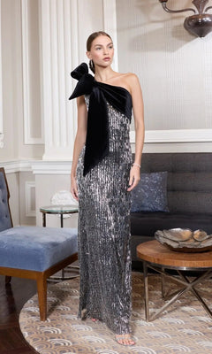 A woman stands in a stylish living room wearing the Mac Duggal 27690 long formal dress in black silver, featuring a one-shoulder design with sequins and a large bow.