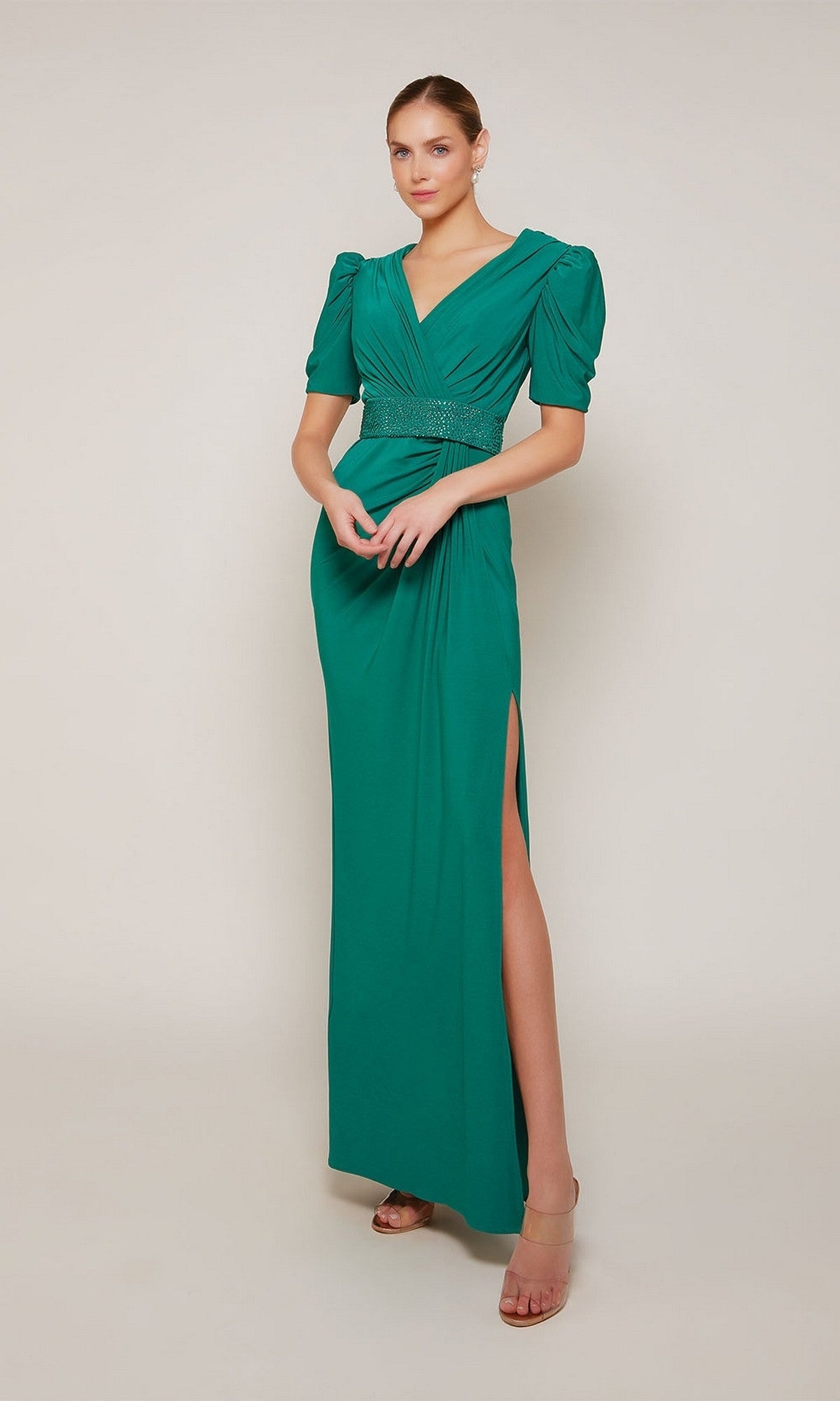 A woman stands against a plain background wearing the Alyce 27690, a long emerald formal jersey dress with puffed short sleeves, V-neckline, beaded belt, and thigh-high slit.