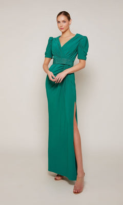 A woman stands against a plain background wearing the Alyce 27690, a long emerald formal jersey dress with puffed short sleeves, V-neckline, beaded belt, and thigh-high slit.