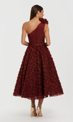 A woman faces away, wearing the Mac Duggal 27698 Midi Wine Wedding Guest Dress with a textured floral skirt and gold heels, set against a plain white background.