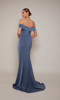A woman in the Alyce 27698 long formal dress—a fitted, off-the-shoulder blue gown—stands facing away with her hands behind her back against a plain background.