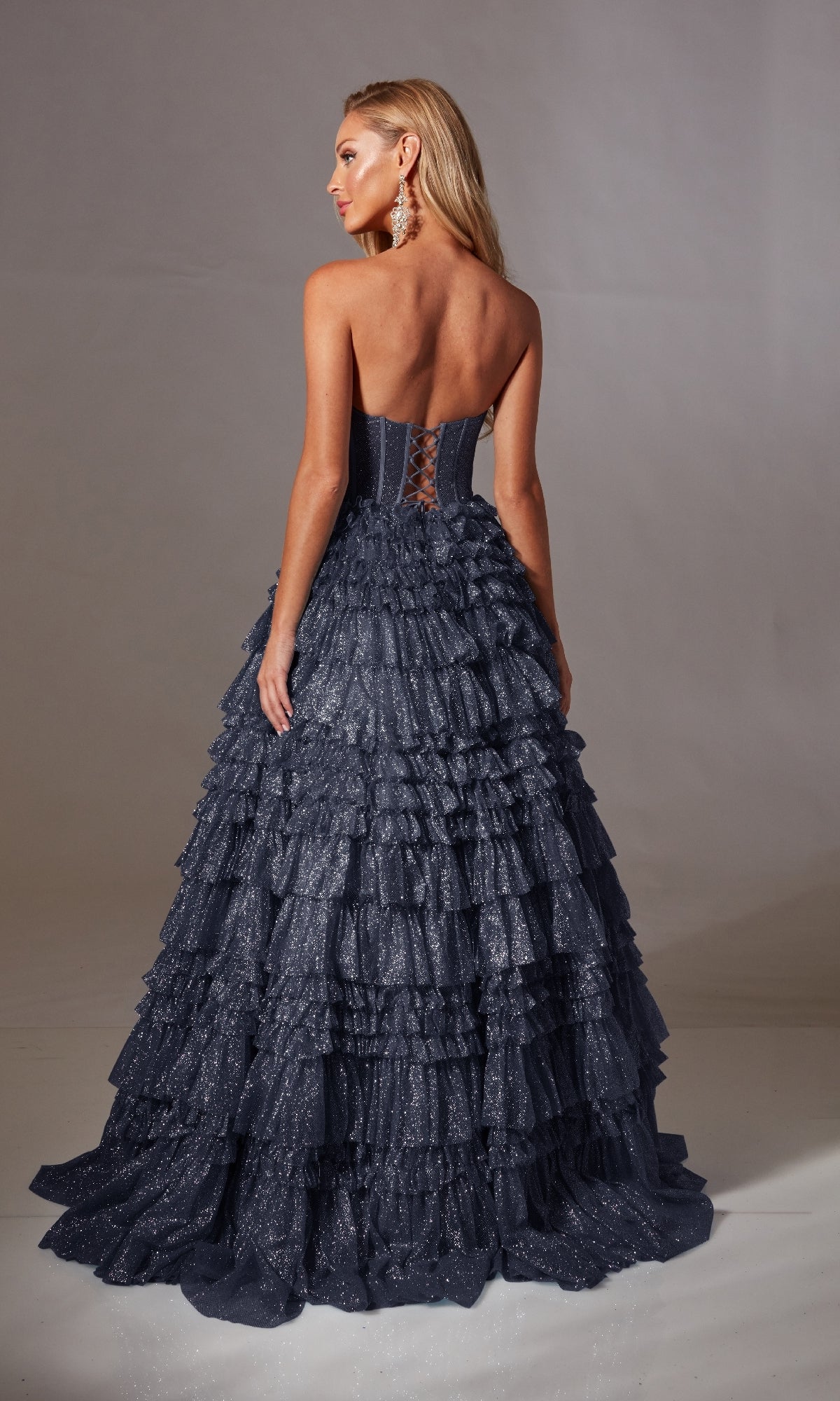 A woman models the Strapless Long Prom Ball Gown: Aerie Couture 276—a sparkly dark blue, strapless, ruffled a-line evening dress with a corset back—standing against a plain background, facing away from the camera.