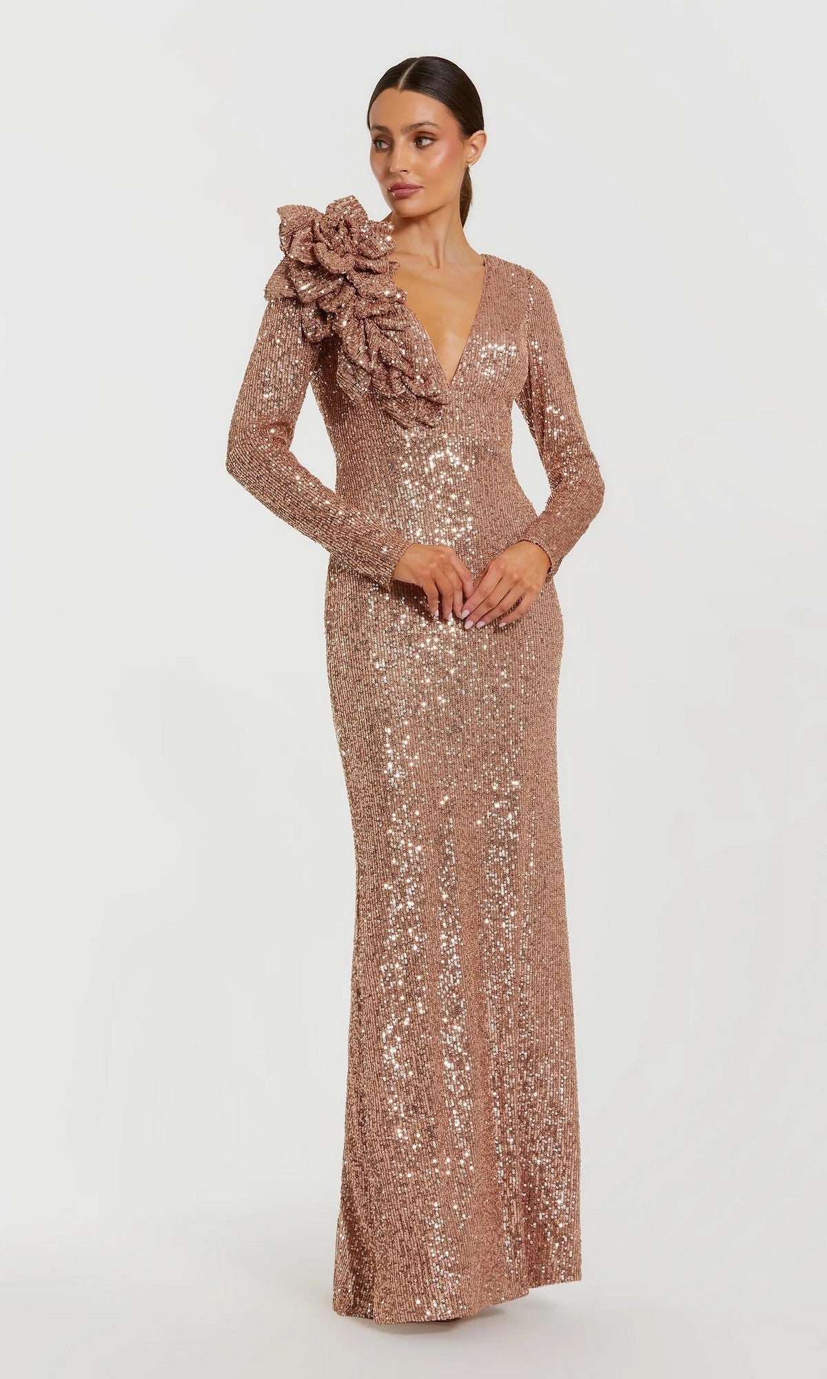 A woman wears the Mac Duggal 27700 long formal dress, featuring sequins, long sleeves, and a striking 3D floral shoulder ruffle, standing against a plain white background.