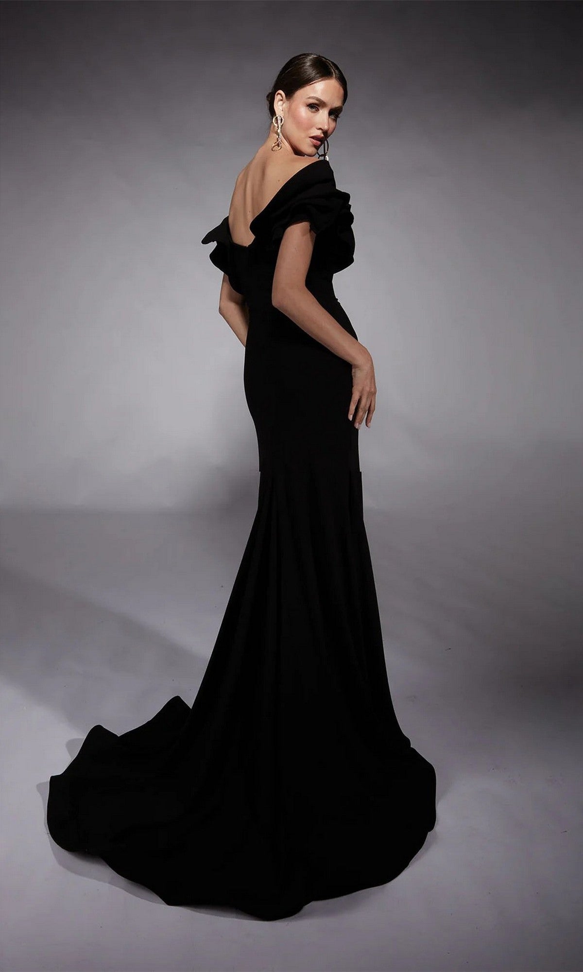 A woman models the Alyce 27714 long formal dress—a black sheath gown with a train, ruffled sleeves, and open back—posing in a studio against a gray backdrop.