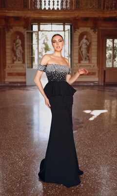 Wearing the Alyce 27715, a striking black off-the-shoulder formal dress with a peplum bodice, a woman poses elegantly in an opulent hall with marble floors and classical statues.