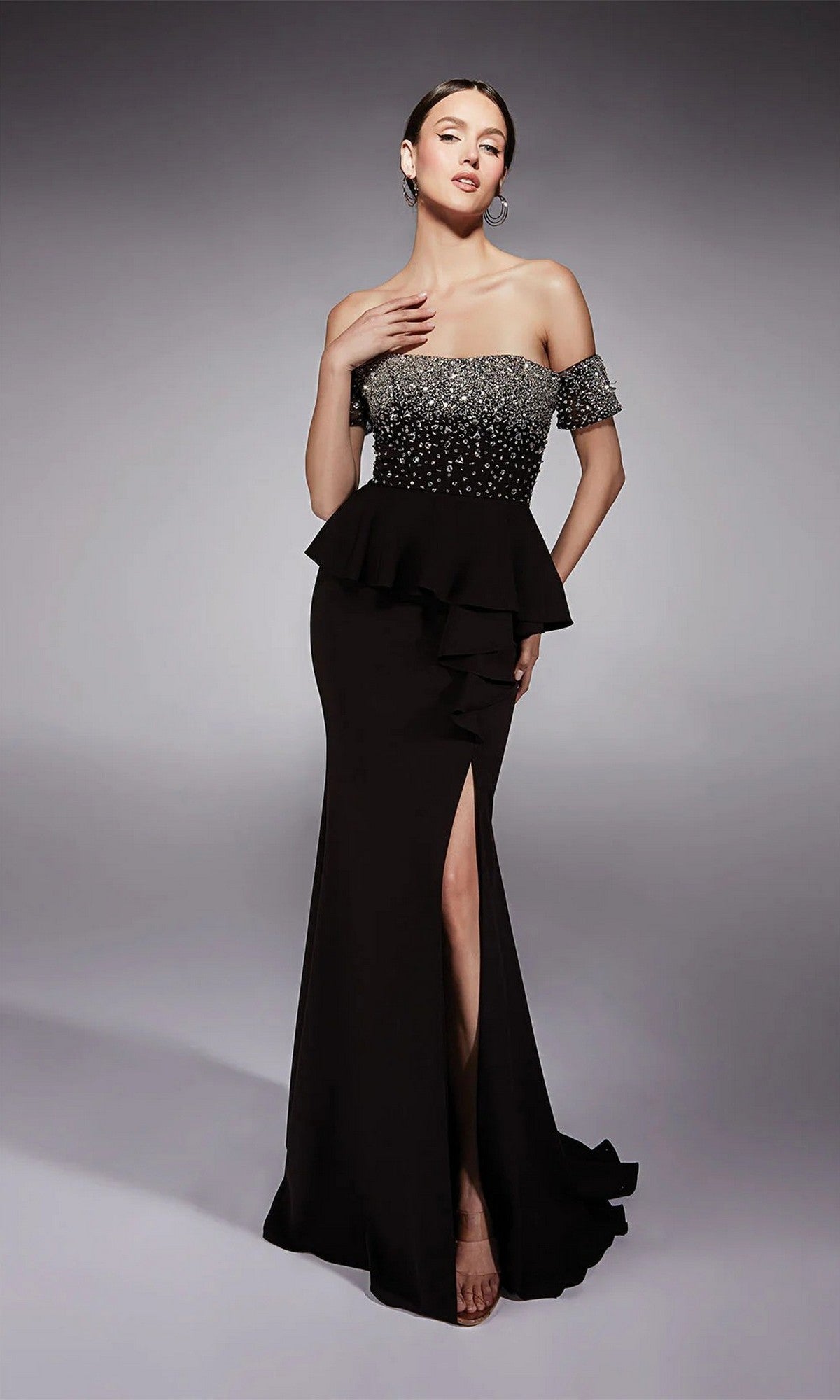 A woman poses in the Alyce 27715 long formal dress in Black/Silver, showcasing a beaded peplum bodice, fit and flare silhouette, and high leg slit against a plain gray background.