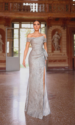 A woman wears the Alyce 27717 long formal dress, featuring textured jacquard fabric, an asymmetrical neckline, and a thigh-high slit, standing in an ornate room with classical statues and open doors in the background.