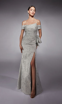 A woman models the silver Long Formal Dress: Alyce 27717, featuring textured jacquard fabric, an asymmetrical neckline, a thigh-high side slit, and a side bow detail, set against a plain gray background.
