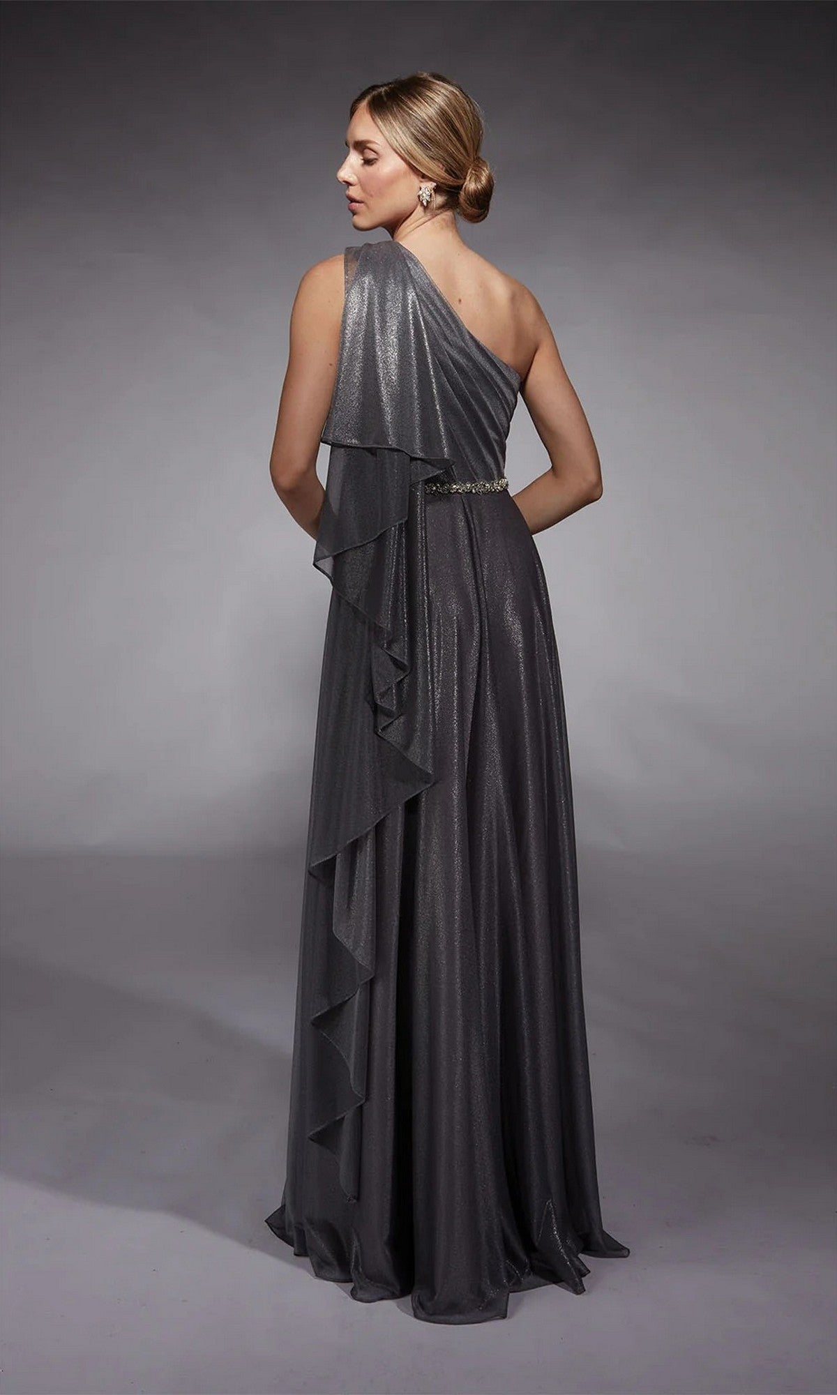 A woman stands against a plain gray background wearing the Alyce 27719, a long, shimmery dark gray one-shoulder formal dress with ruffled detailing down the back.