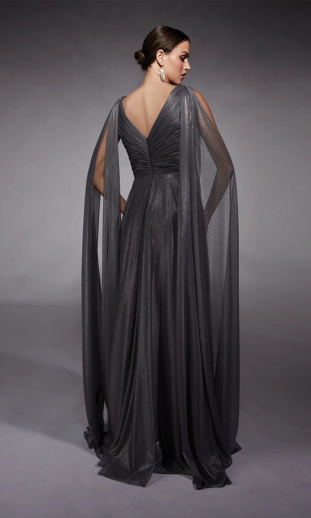 A woman stands against a gray background, facing away in the Alyce 27720—an elegant floor-length dark gray metallic chiffon gown with draped sheer cape sleeves, perfect for any mother-of-the-bride moment.