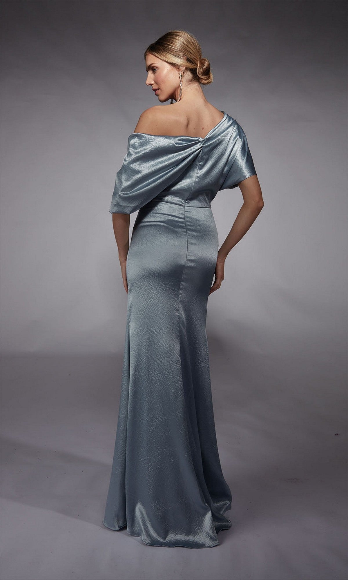 A woman models the Alyce 27721 Long Formal Dress—a silver, off-the-shoulder sheath gown—standing with her back turned and glancing over her shoulder against a plain gray backdrop.