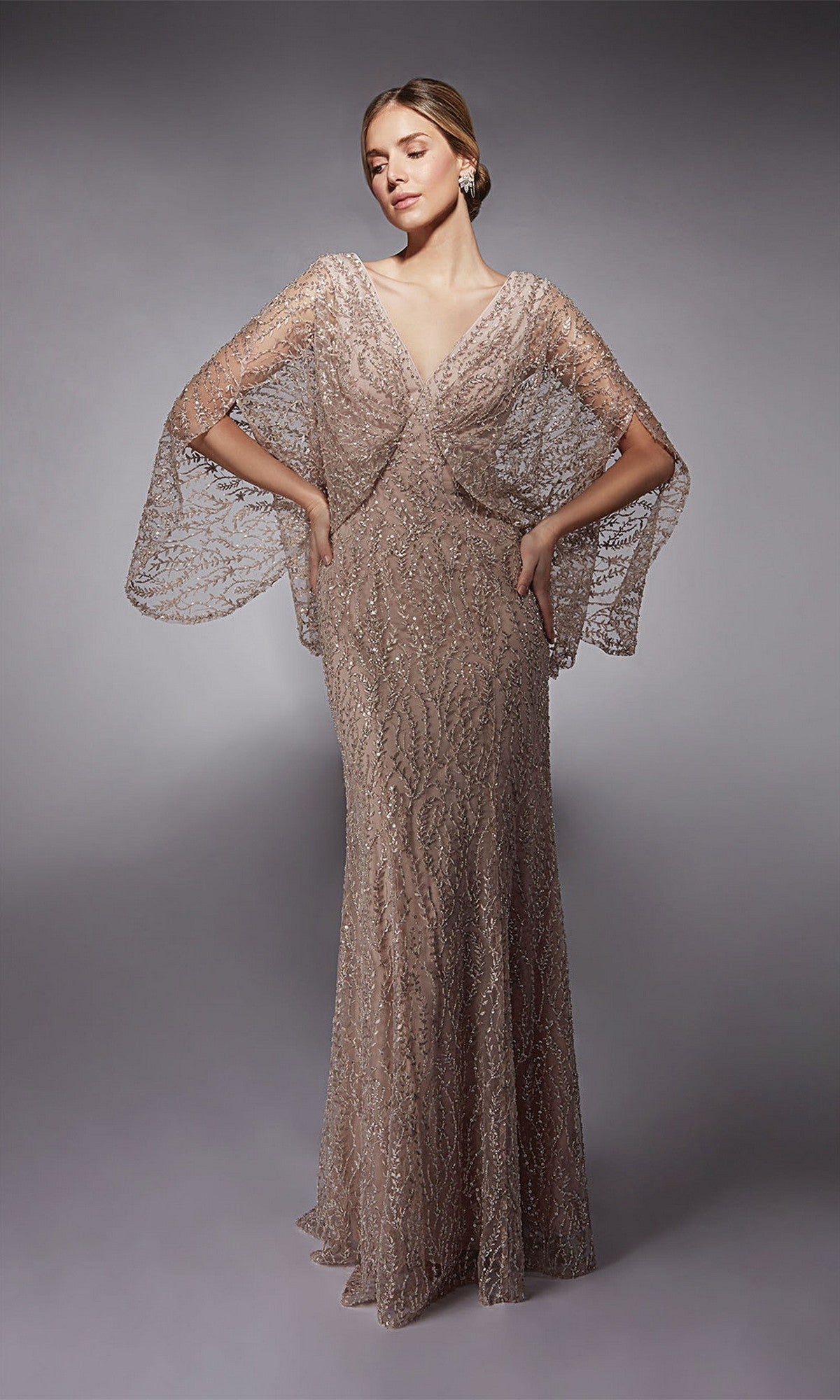 A woman stands with hands on hips against a plain gray background, wearing the Alyce 27724 long formal dress in sultry blush, featuring sheer beaded cape sleeves and crystal embellishments.