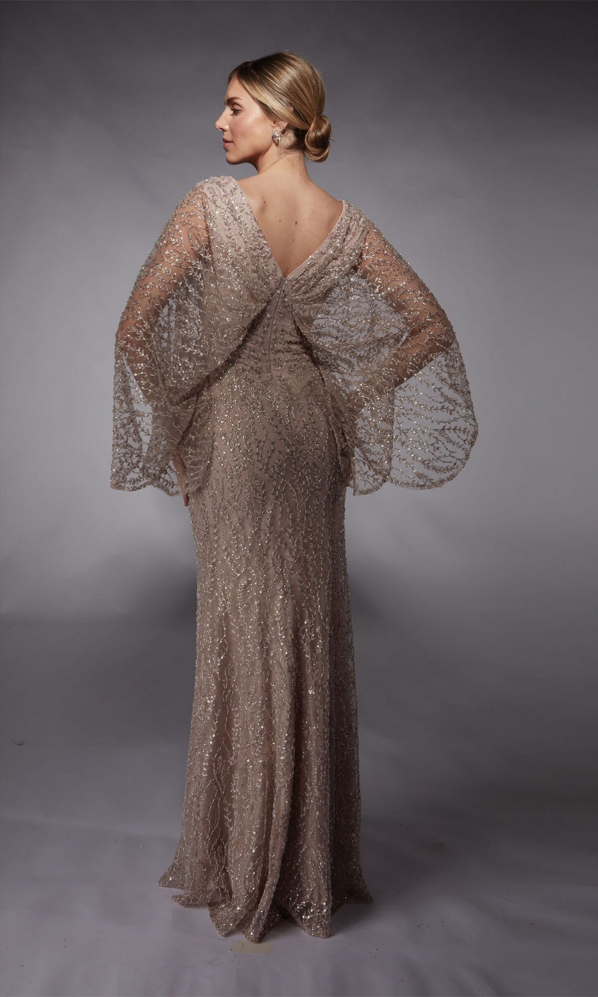 A woman stands in a studio, facing slightly away, wearing the Alyce 27724 Long Formal Dress—a shimmering, floor-length gown with crystal beaded embellishments, sheer wide sleeves, and a V-shaped back.