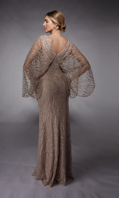 A woman stands in a studio, facing slightly away, wearing the Alyce 27724 Long Formal Dress—a shimmering, floor-length gown with crystal beaded embellishments, sheer wide sleeves, and a V-shaped back.