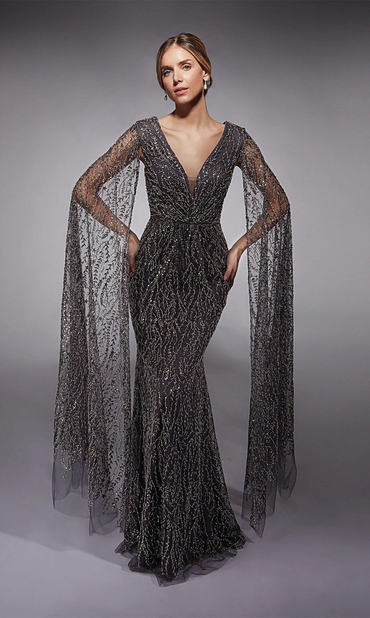 A woman in the Alyce 27725 long formal dress in charcoal, featuring intricate beading, a fitted sheath silhouette, and sheer cape-like sleeves, poses against a neutral background.
