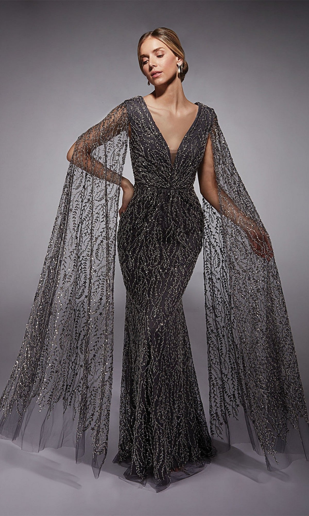 A woman poses against a neutral background wearing the Alyce 27725 long formal dress—an elegant, fitted sheath gown with intricate metallic beadwork and dramatic sheer cape sleeves.
