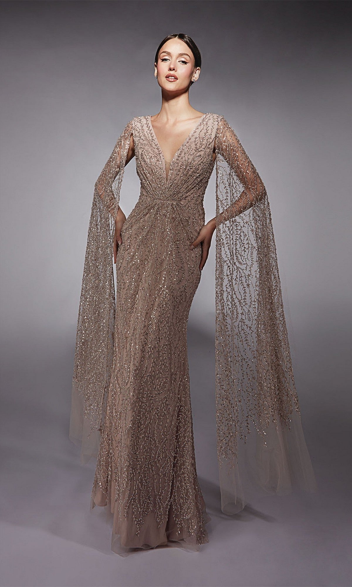 A woman poses elegantly in the Alyce 27725 Long Formal Dress in Sultry Blush, a shimmering sequin evening gown with sheer beaded cape sleeves and a fitted sheath silhouette, set against a plain gray background.