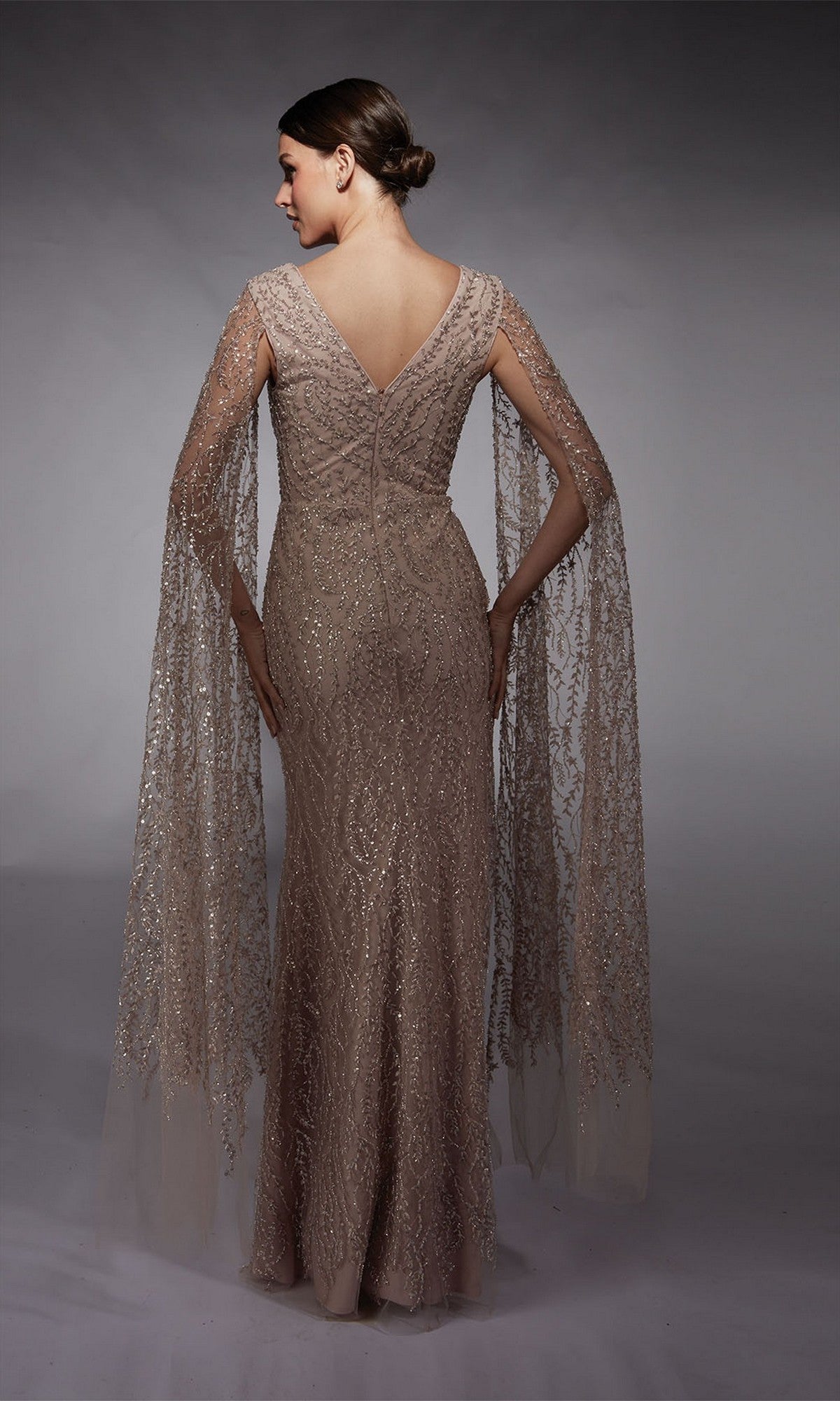 A woman in the Alyce 27725 long formal dress—an embellished floor-length sheath with sheer, beaded cape sleeves—stands with her back to a gray background.