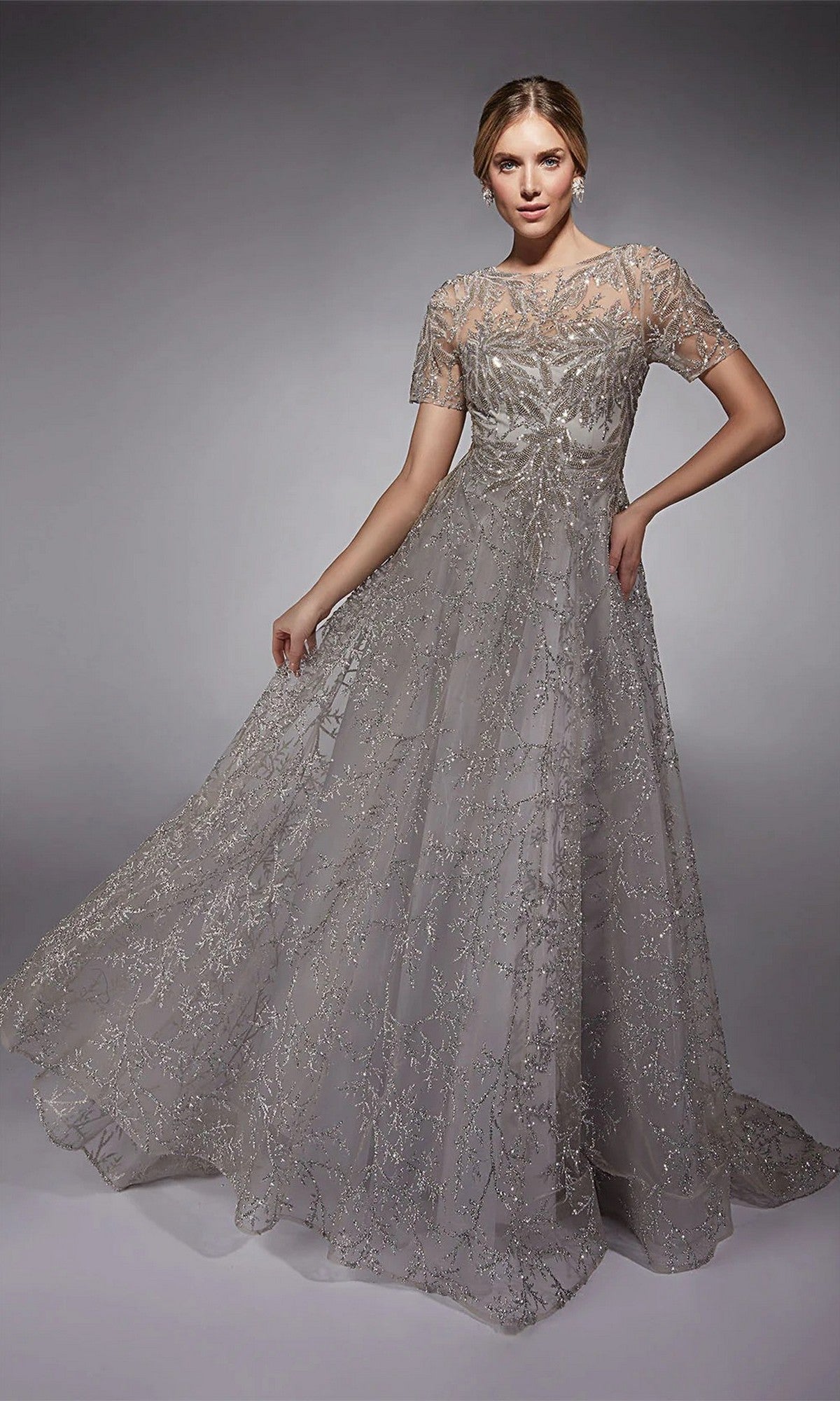 A woman poses against a plain gray background in the Alyce 27726 long formal dress, featuring short sleeves, intricate beaded details, and shimmering silver glitter tulle.