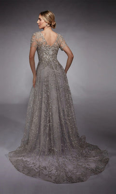 A woman faces away in the Alyce 27726 long formal dress—a silver, floor-length gown with intricate beadwork, glitter tulle, and a V-back—set against a gray studio backdrop.