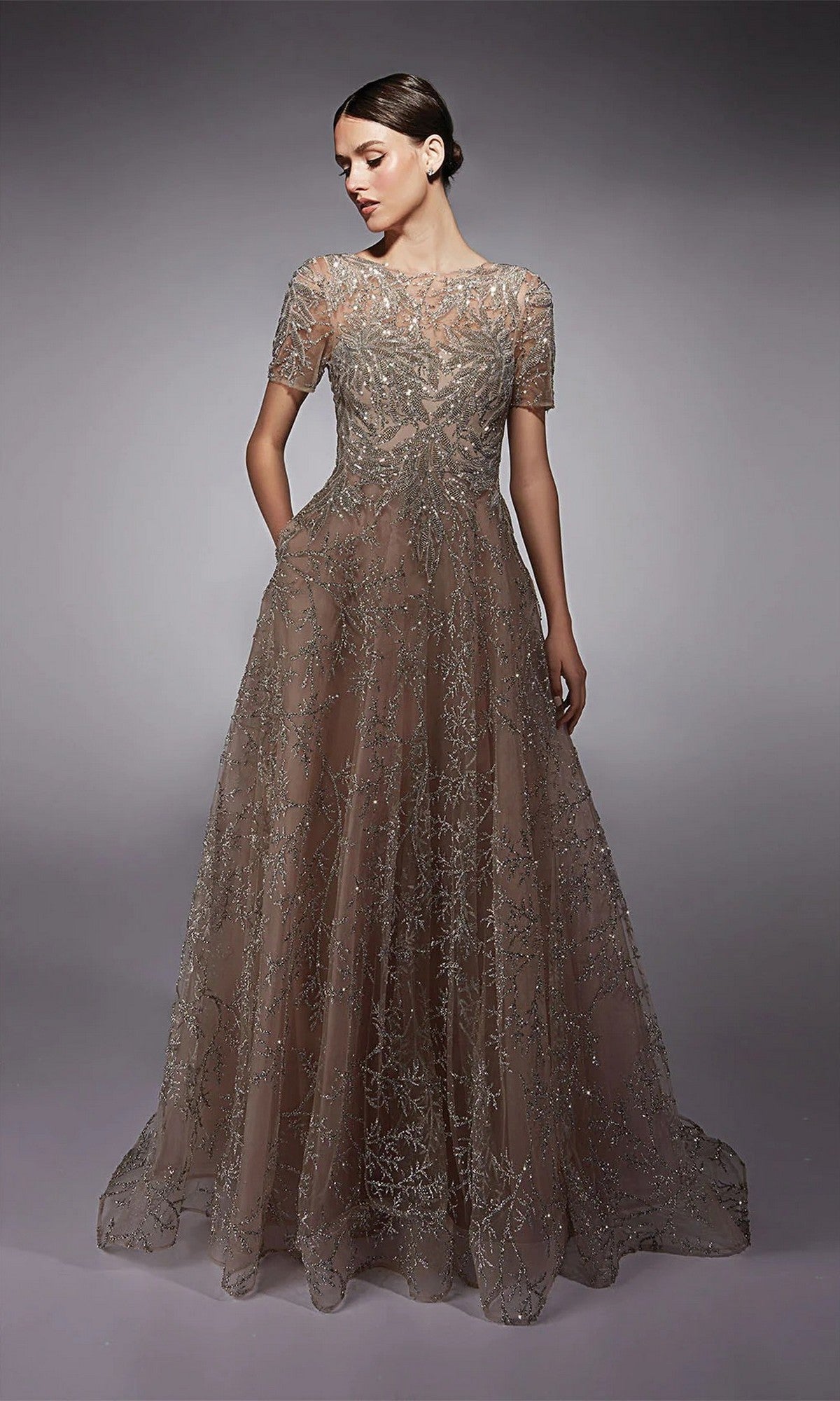 A woman in the Alyce 27726 long formal dress in Cashmere Rose, featuring short sleeves, intricate silver beadwork, and glitter tulle, stands against a plain gray background.