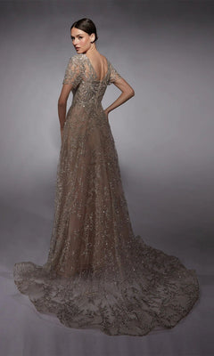 A woman poses in the Alyce 27726 long formal dress, showcasing short sleeves, a V-back, and a sheer embroidered glitter tulle train against a gray studio backdrop.