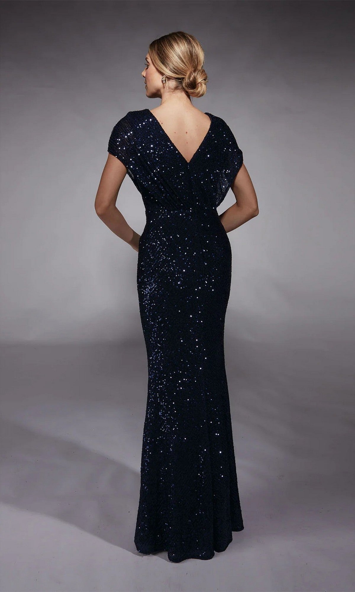A woman with blonde hair in a low bun wears the Alyce 27729, a floor-length, short-sleeved dark blue sequin mother-of-the-bride gown with a V-back, standing against a plain background.