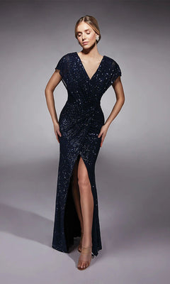 A woman poses in a studio wearing the Long Formal Dress: Alyce 27729, a dark blue sequin mother-of-the-bride gown with a V-neck, short sleeves, and front slit. Her hair is styled back and she completes the look with nude heels.