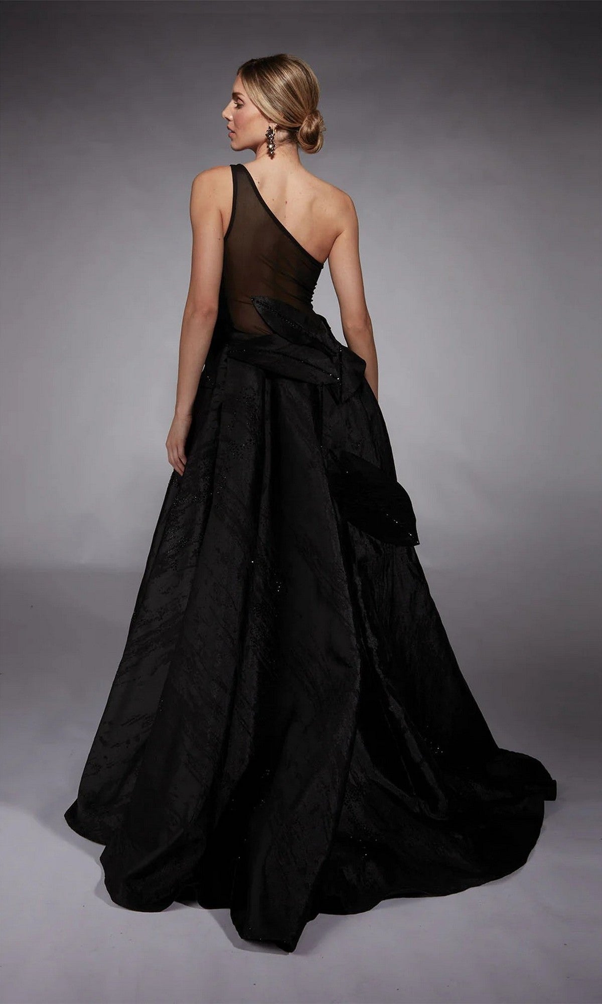 A woman stands with her back to the camera, showcasing the Alyce 27730 long formal dress—a black one-shoulder gown with a sheer back panel and voluminous skirt—an elegant choice for today's mother-of-the-bride.