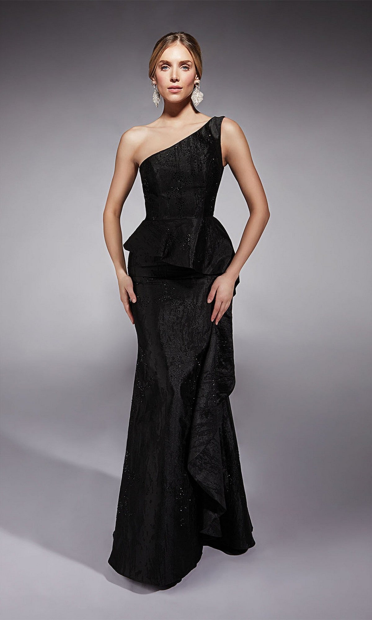 A woman stands confidently against a plain gray background wearing the Alyce 27732 long black formal dress—a jacquard one-shoulder gown with a peplum waist.