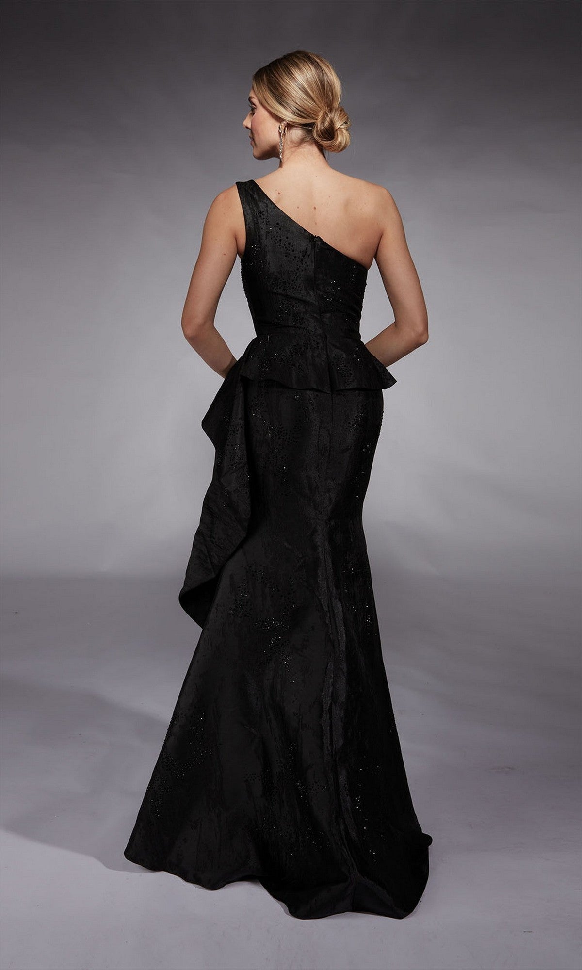 A woman with blonde hair in a low bun wears the Alyce 27732 Long Formal Dress—a black, one-shoulder, floor-length gown with a fitted bodice and flared skirt—while standing with her back to the camera.