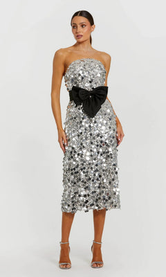 A woman models the Mac Duggal 27735 midi semi-formal dress with silver sequins, a strapless neckline, black waist bow, and silver high-heeled sandals against a plain light background.
