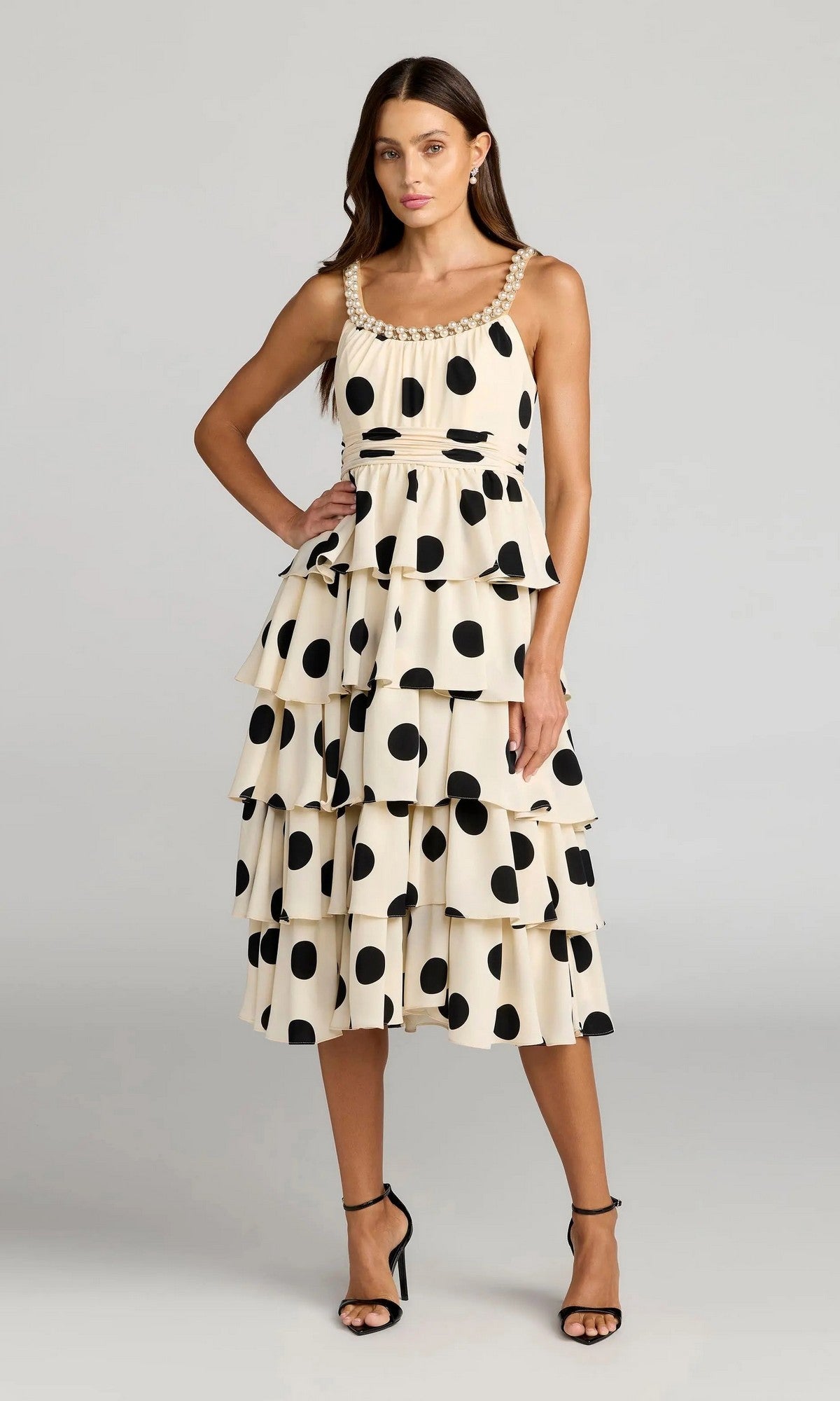 A woman wears the Mac Duggal 27736 Midi Party Dress in Black Ivory, showcasing its sleeveless, tiered silhouette with bold black polka dots, and completes her playful look with strappy sandals as she stands against a plain gray background.