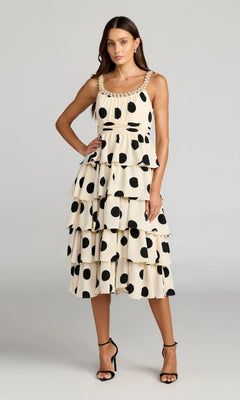 A woman wears the Mac Duggal 27736 Midi Party Dress in Black Ivory, showcasing its sleeveless, tiered silhouette with bold black polka dots, and completes her playful look with strappy sandals as she stands against a plain gray background.
