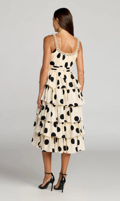 A woman stands with her back to the camera, wearing the Mac Duggal 27736 Midi Party Dress—an ivory, tiered party dress adorned with large black polka dots—paired with black high heels.