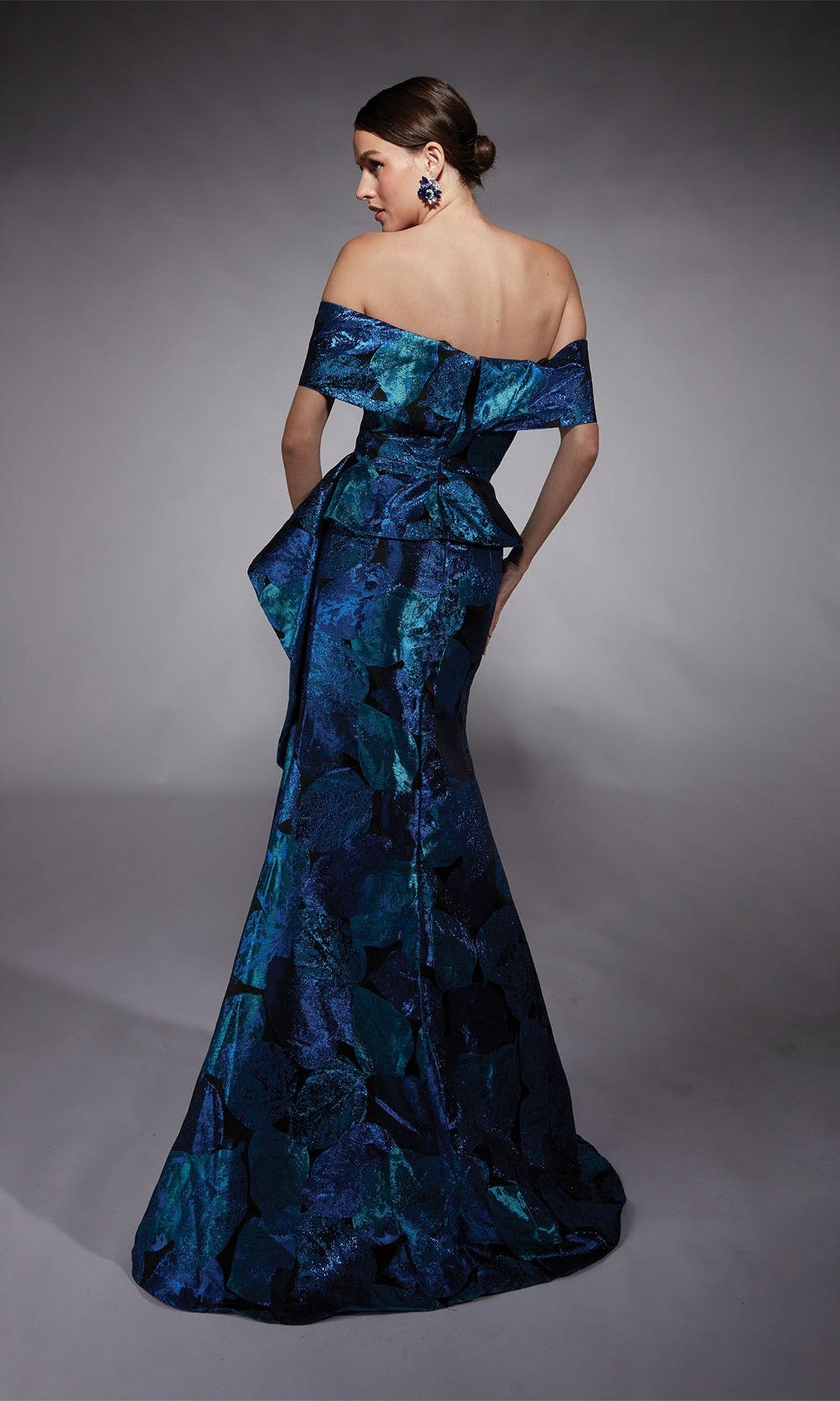 Woman poses with her back turned in a studio, wearing the Alyce 27736 long formal dress: an off-the-shoulder, floor-length blue and green patterned gown with a fitted waist and fit-and-flare silhouette.