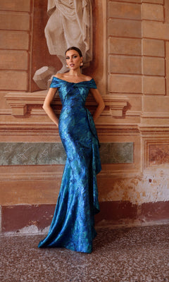 A woman in the Alyce 27737 long formal dress, featuring a blue off-the-shoulder design, stands against a textured wall with a classical relief sculpture in the background.