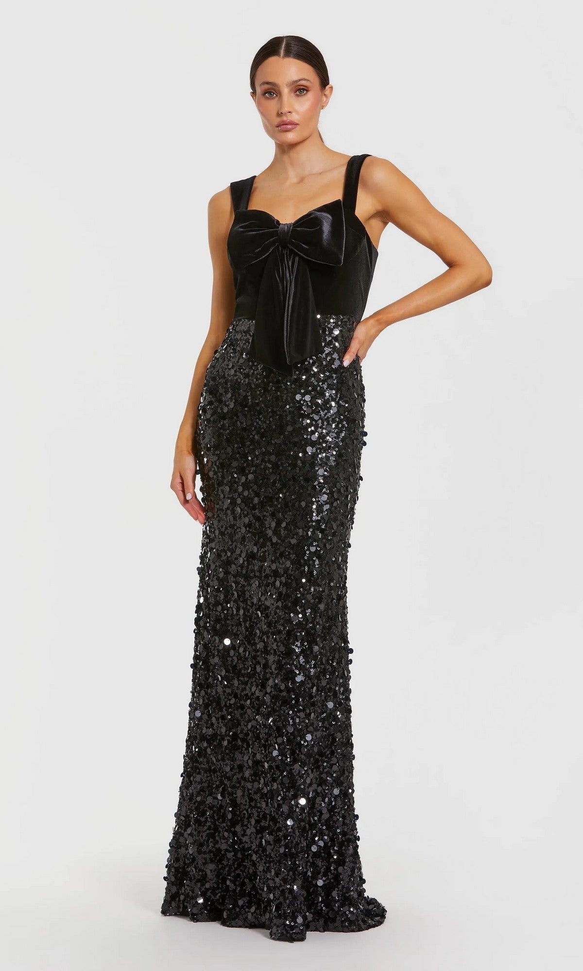 A woman models the Mac Duggal 27738 long formal dress in black, with a velvet bodice, large bow accent, and a sequined floor-length skirt, posed before a plain light backdrop.