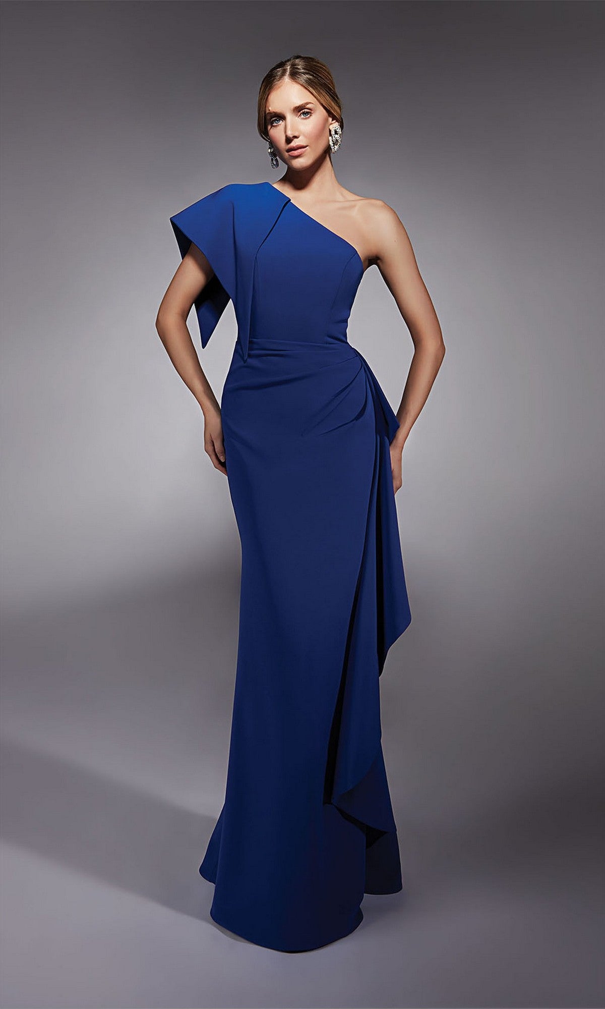 A woman poses in the Alyce 27738 long formal dress, featuring a floor-length, one-shoulder design with draped detailing and statement earrings, set against a gray background.