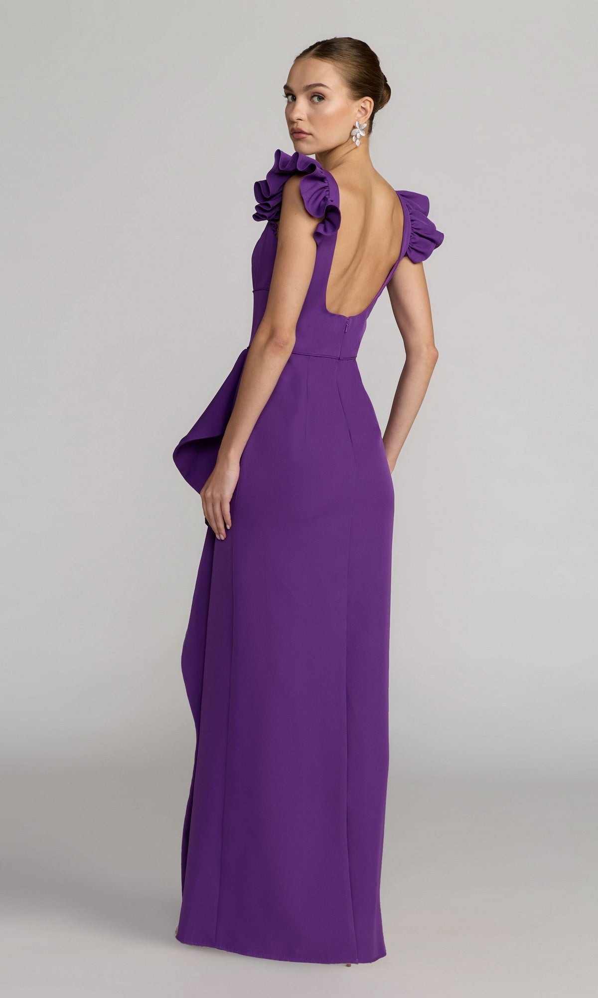 A woman stands in a studio setting, looking over her shoulder, wearing the Mac Duggal 27740 long formal dress—a floor-length, sleeveless purple gown featuring ruffled shoulders, a square neckline, and a low back.